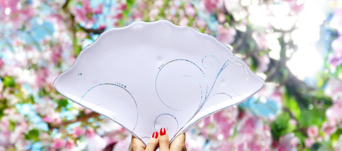 Fan shaped pink charger plate with a floral pattern held up by a woman's hand with cherry blossoms in the background.