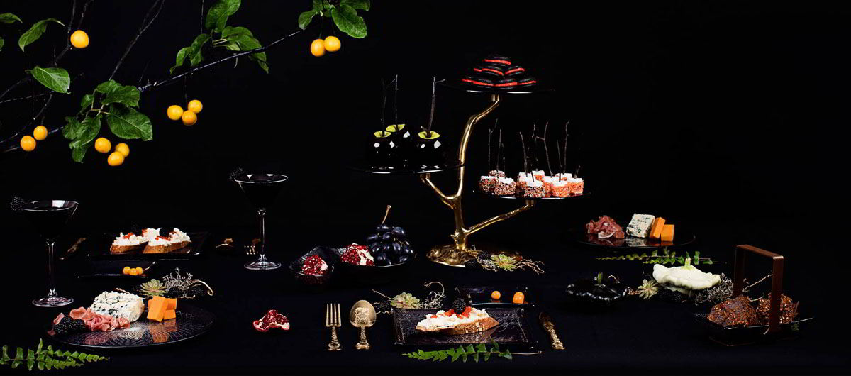 Dark and mysterious Halloween tablescape with a dark blue 3 tiered high tea stand on a bronze tree, twin bowls with chocolate, square plates with floral pattern with appetizers, fruit bowl with fruits inside and finger food platter in Filigree pattern.