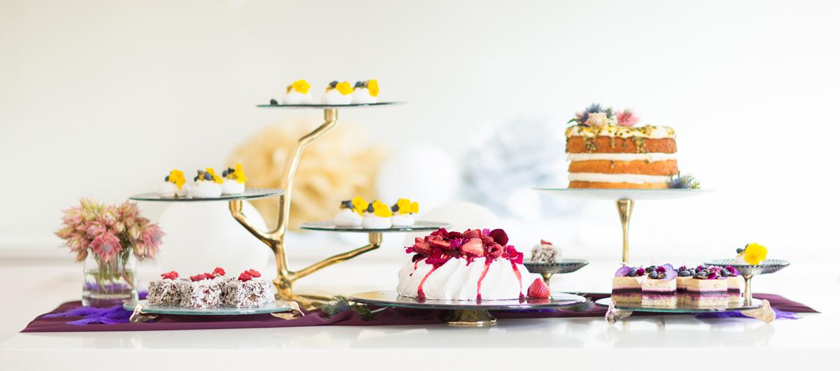 Different luxurious glass cake stands and cake display stands with bronze pedestals with cakes, a Pavlova, lamingtons and petit fours.